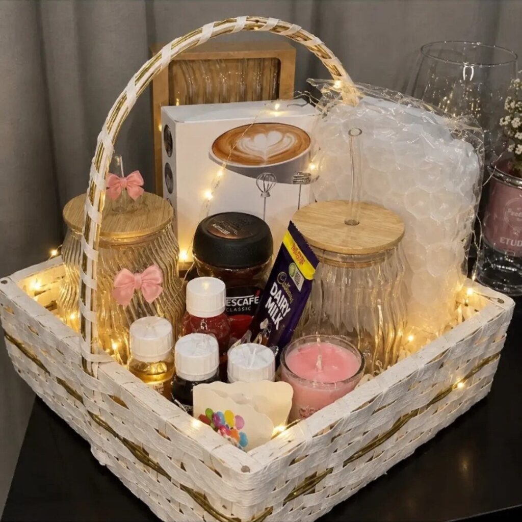 White woven gift basket filled with coffee, chocolate, candles, and warm fairy lights.