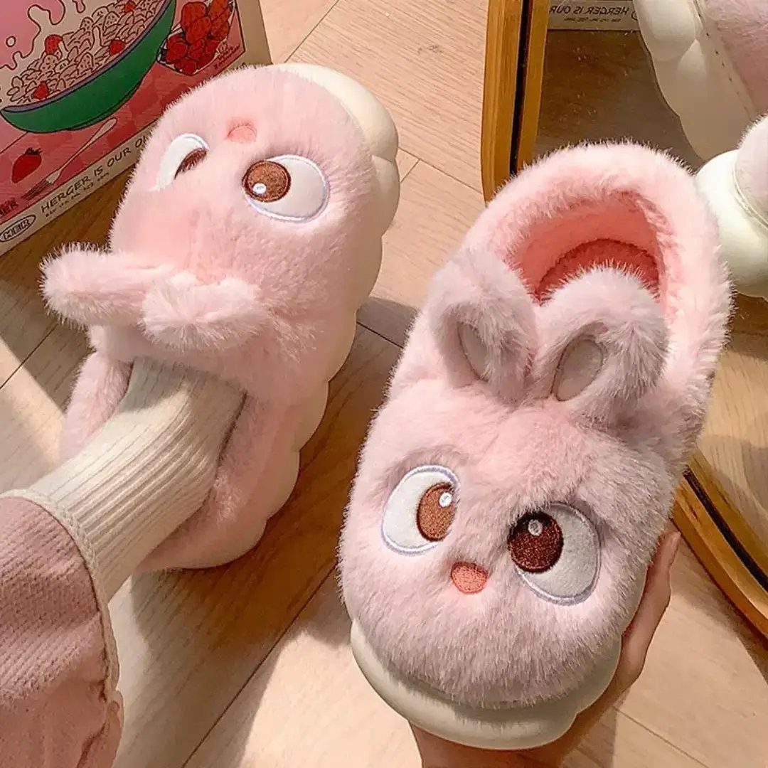 Pink fuzzy plush slippers with cute character faces on a light wood floor.