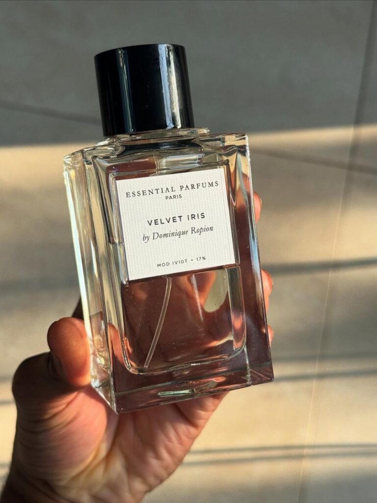 Glass bottle of Velvet Iris cologne held in a hand against a wall