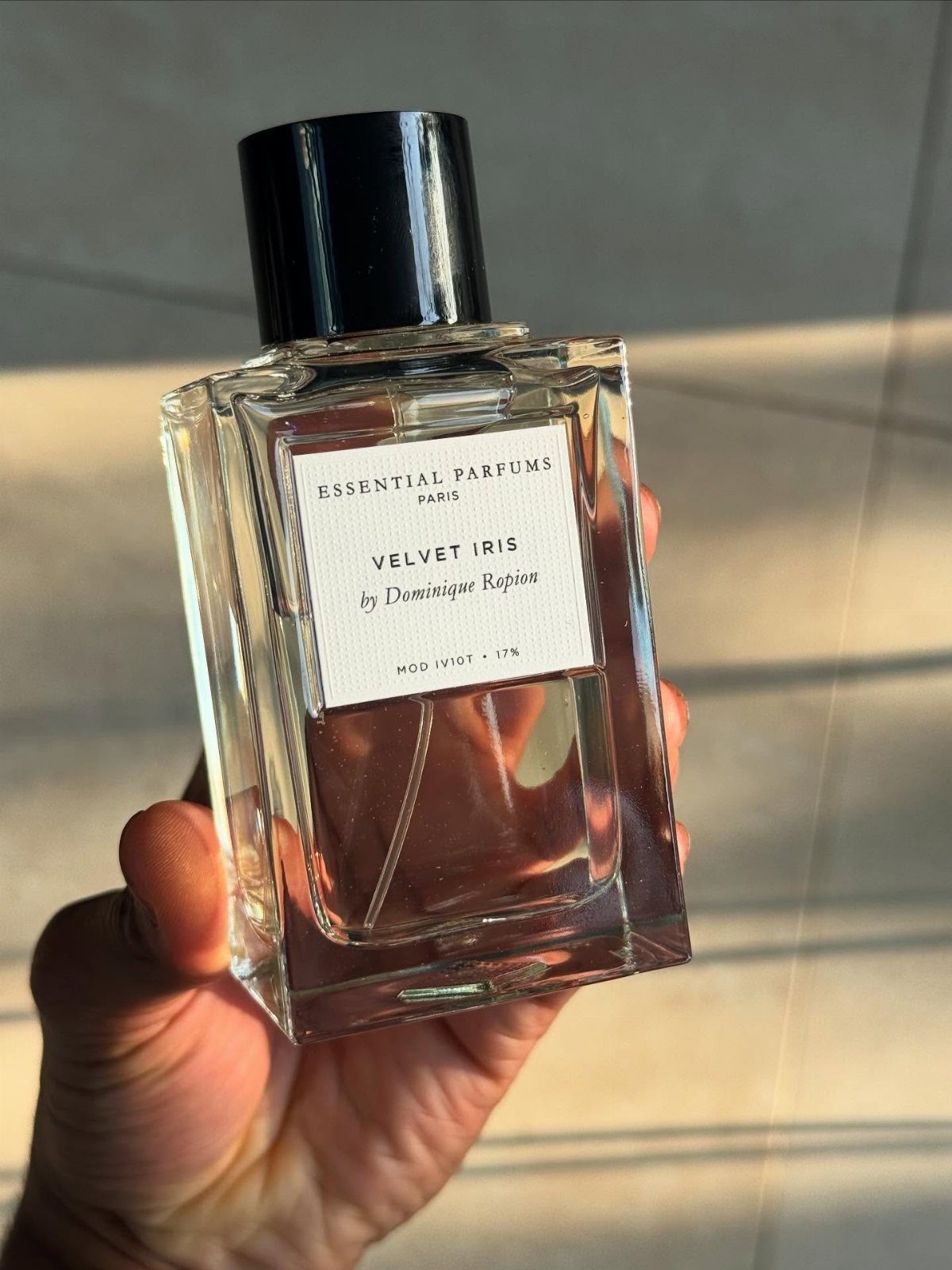 Glass bottle of Velvet Iris cologne held in a hand against a wall