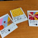 Three music-themed notecards with colorful art and digital music player interfaces on wood.