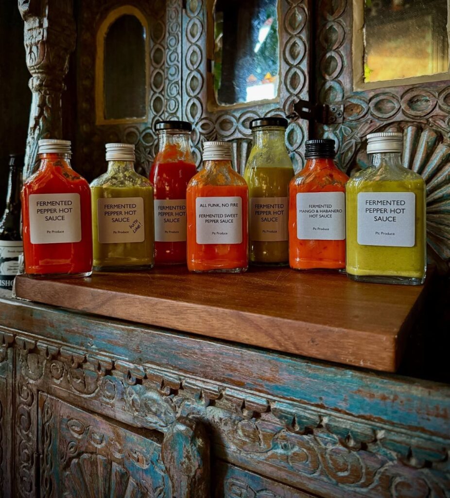 Six colorful bottles of fermented pepper hot sauce on a carved wooden shelf.