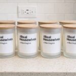 Eight frosted glass candles with wooden lids and personalized labels on a counter.