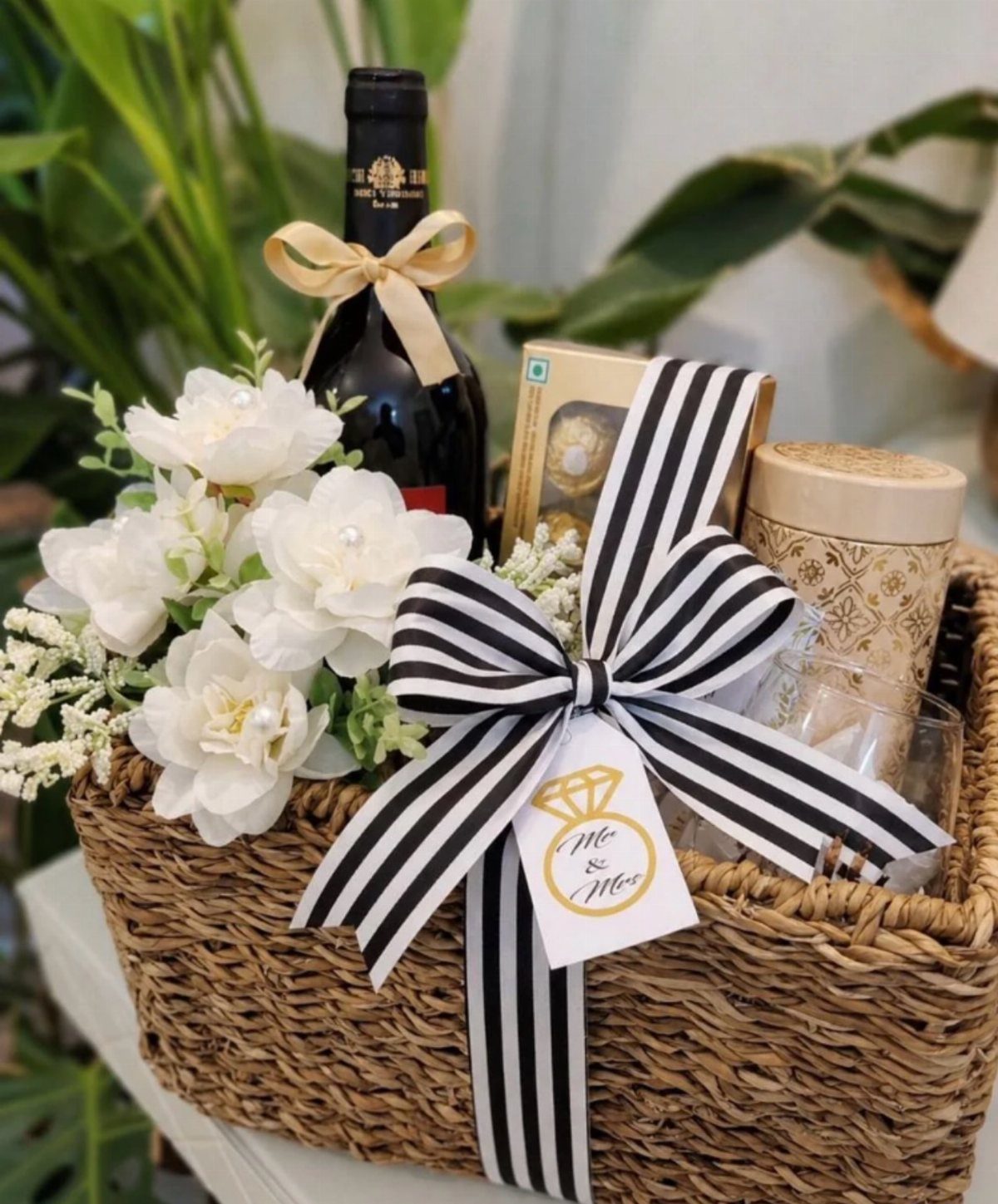 Woven gift basket with red wine, white flowers, and black striped ribbon.